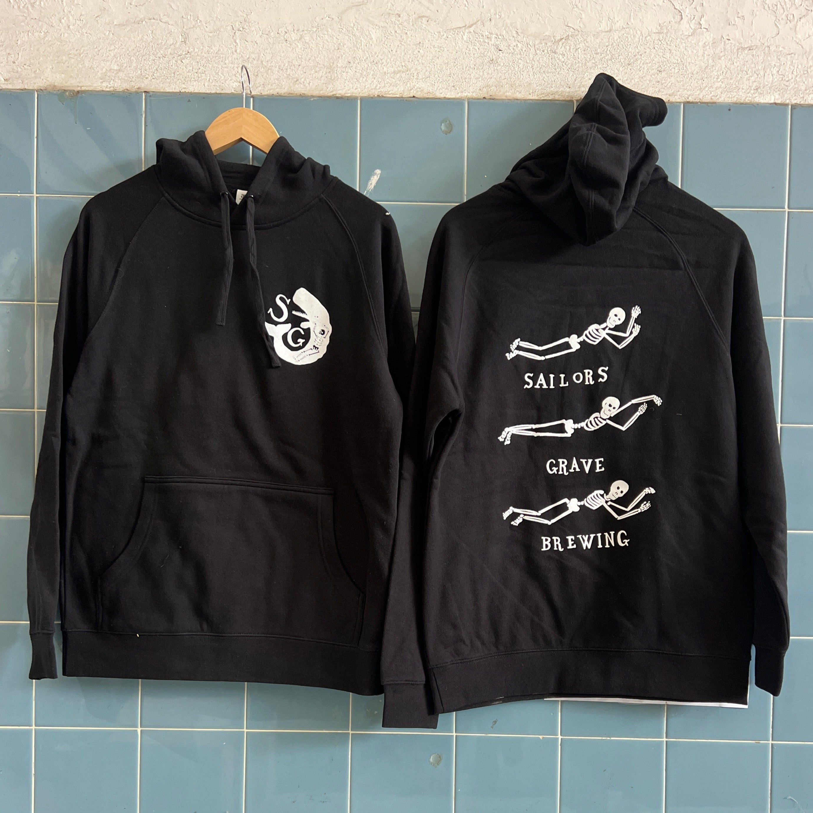 Two black hoodies with white Sailors Grave Brewing logo on the front and back, displayed against a blue-tiled wall.
